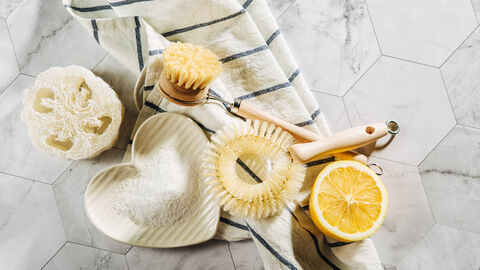 Spülbürsten aus Holz, halbierte Zitrone, Natron in einer Schale, gestreiftes Küchentuch und Luffaschwamm auf gefliesten Boden