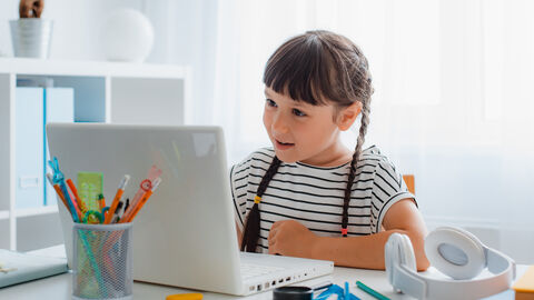 Ein Mädchen sitzt vor eine Notebook, auf dem Tisch liegen verschiedene Schreib- und Schulutensilien