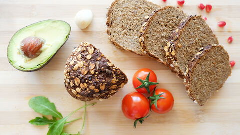 Aufgeschnittenes Breot liegt auf einem Holztisch neben Tomaten, Basilikum und einer halben Avocado