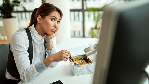 Eine junge Frau sitzt vorm Computer und isst ihr Mittagessen
