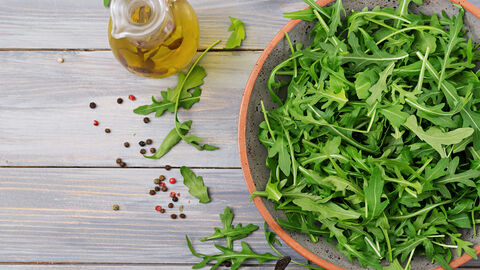 Rucola in einer Schale von oben fotografiert, daneben einige lose Rucolablätter und eine Ölkaraffe