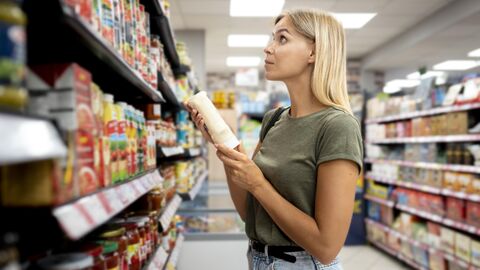 Frau steht im Supermarkt mit einer Mayonnaiseflasche in der Hand und schaut skeptisch ins Lebensmittelregal