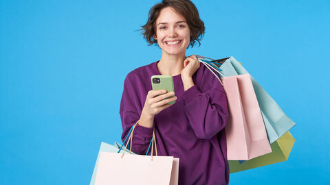 Junge Frau mit kurzer brauner Bob-Frisur hat über die Schulter drei Einkaufstütengeschlagen und hat in der anderem Hand ein Smartphone und ums Handgelenk eine weitere Einkaufstüte.