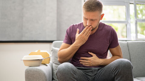 Mann sitzt auf Sofa und hält sich den Bauch. Er macht ein schmerzverzerrtes Gesicht, muss aufstoßen und hält sich die andere Hand vor den Mund. Auf der Sofale stehen Plastik-To-go-Verpackungen für Lebensmittel.