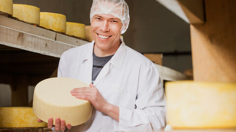 Mann mit weißem Kittel und Haube auf dem Kopf hält einen Laib Käse in der Hand. Im Hintergrund sind Holzregale mit weiteren Laiben Käse.