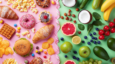 Auf der rechten Seite liegen unterschiedliche Süßigkeiten (Donut, Waffel, Berliner, Croissant, Chips), auf der rechten Seite liegen unterschiedliche Obst- und Gemüsesorten (Cocosnuss, Äpfel, Gurke, Tomaten, Zucchini, Banane, Blutorange, Blaubeeren, Trauben). In der Mittel liegt ein Maßband.