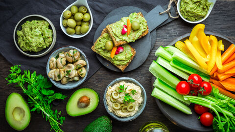 Verschiedene Rohkostsnacks in Schälchen, zum Beispiel Oliven, Gemüsesticks aus Paprika, Gurke und Sellerie, Tomaten, eingelegte Pilze, Avocadocreme, ein paar Stängel Petersilie.