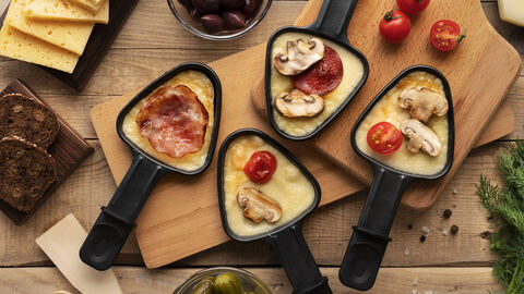 Vier Raclettepfännchen von oben mit Käse, tomate, Salami, Pilzen und schinken. Drum herum stehen Schälchen mit weiteren Raclettezutaten wie saure Gurken, Oliven, Brot und Käse.