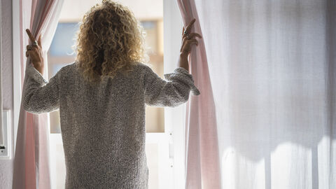Eine Frau steht mit dem Rücken zum Fotografierenden am Fenster und zieht die Vorhänge ein Stück auseinander, um aus dem Fenster zu sehen.