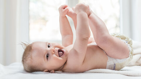 Baby in Windel liegt auf Rücken, gfreift sich an die Füße und lacht