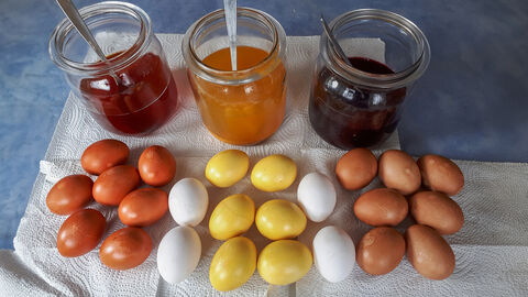 Drei Gläser mit Farblösung und davor die damit gefärbten Eier in goldbraun, gelb und rotbraun