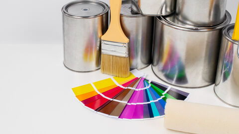 Verschiedene Farbdosen gestapelt, Pinsel und Farbrolle sowie ein Farbfächer liegen auf einer hellen Fläche