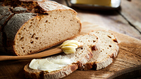 Ein aufgeschnittenes Roggenbrot liegt auf einem Holzbrett. Auf der ersten Brotscheibe liegt ein Holzlöffel mit Butter drauf.