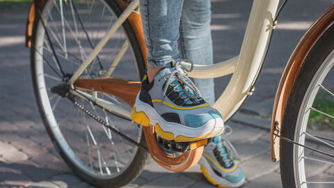 Eine Frau setht mit einem Fuß auf der Fahrradpedale. Außerdem sieht man in dem Bildausschnitt noch das Hinterrad und einen Teil des Fahrradrahmens.