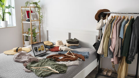 Neben einem Bett steht eine Kleiderstange auf der Kleidung hängt, zum Beispiel Jacken, Kleider und Hemden und ein Hut. Auf dem Bett liegt ein aufgeklappter Laptop und weitere Kleidung verstreut sowie Kleiderbügel. Auf dder anderen Seite des Bettes steht ein Regal mit einigen Pflanzen.