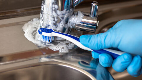 Jemand in Nitrilhandschuhen putzt mit einer Zahnbürste den Wasserhahn eines Waschbeckens, um den Kalk zu entfernen