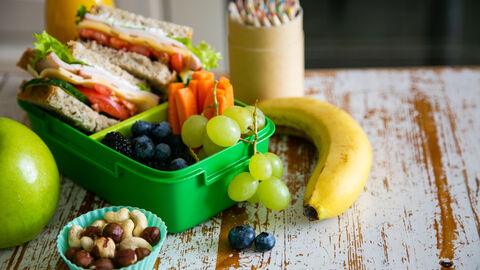 Bortdose mit belegten Broten mit Käse, Tomaten und Salat, Heidelbeeren, Weintrauben und Möhrensticks. Daneben steht ein kleines Schälchen mit Nüsse. Links und rechts liegen ein Apfel und eine Banane.