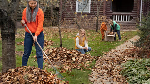 Eine Frau kehrt im Garten Laub zusammen. Im Hintergrund sind Kinder zu sehen die dabei helfen