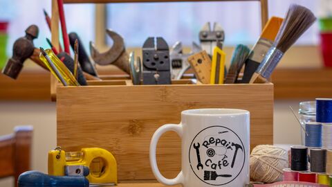 Kaffeetasse mit Repair Cafe-Aufschrift und viel Materialien und Werkzeug für Reparaturen von  Alltagssachen