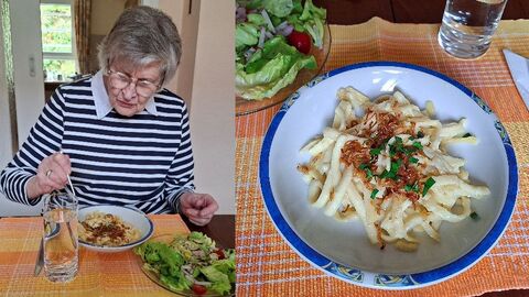 Seniorin sitzt am Esstisch und isst. Vor ihr steht ein Teller mit Käsespätzle und ein kleinerer Teller mit einem Beilagensalat. 