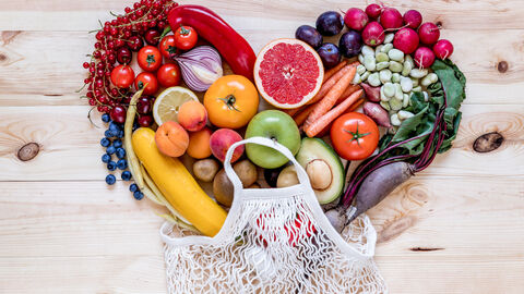 Auf einem Holztisch liegt verschiedenes buntes Obst und Gemüse zu einem Herz geformt. An der Herzspitze ist eine gehäkelte Einkaufstasche drapiert, sodass es aussieht als würde das Herz aus der Einkaufstasche kommen.