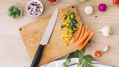 Blick von oben auf ein Holzschneidebrett. Auf dem Brett liegt ein Küchenmesser sowie kleingeschnittene Paprika und Möhren. um das Schneidebrett herum stehen kleine Schälchen mit kleingeschnittenen Zwiebeln und ebenfalls Paprika. Zudem liegen vereinzelt Pilze und Radieschen um das Schneidebrett herum.