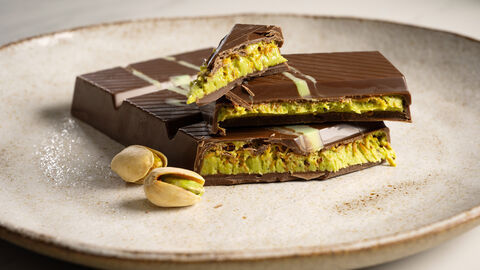 Eine zerbrochene Tafel Dubai-Schokolade auf einem Teller. Daneben liegen Pistazien.