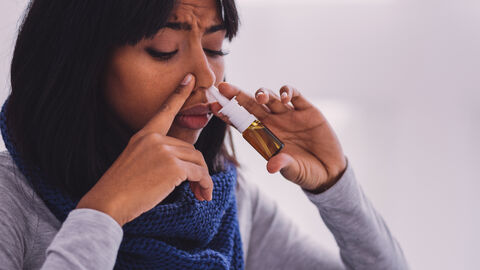 Eine Frau sprüht sich ins linke Nasenloch Nasespray und hält mit der rechten Hand das rechte Nasenloch zu.