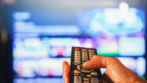 Eine Hand hält eine Fernbedienung in der Hand und richtet diese auf einen Fernseher. 