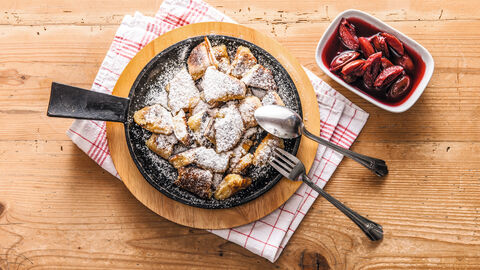 Blick von oben auf eine Pfanne mit Kaiserschmarrn mit Puderzucker. an der Pfanne lehnt rechts das Besteck. Die Pfanne steht auf einem Holzbrett, das wiederum auf einem rot-weißem Geschirrtuch steht. Neben der Pfanne befindet sich ein Schälchen mit Zwetschgenröster.
