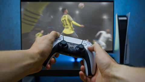 Man sieht einen Fernseher und daneben eine Playstation-Konsole. Auf dem Bildschirm sieht man ein Fußball-Onlinespiel. Im Vordergrund sieht man zwei Hände, die einen Controller halten.