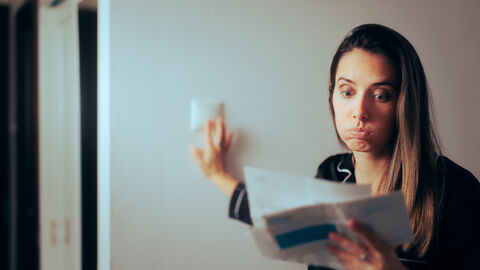 Hohe Energiekosten Eine Frau schaut auf einen geöffneten Brief und macht ein prustendes, besorgtes Gesicht. Mit der rechten Hand betätigt sie einen Lichtschalter.