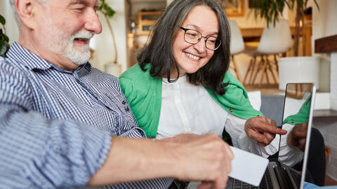 Ein älteres Pärchen sitzt im wohnzimmer vor dem Laptop und schautr gemeinsam auf den Bildschirm. Der Mann hält in einer Hand eine Geldkarte. Die Frau deutet mit dem Finger auf den Bildschirm.