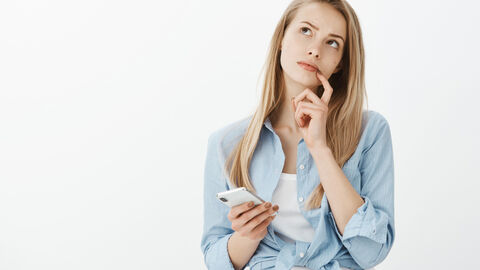 Eine junge Frau hält ein Smartphone in der Hand und schaut fragend nach oben. Mit dem Zeigefinger ihrer linken Hand berührt sie ihren linken Mundwinkel