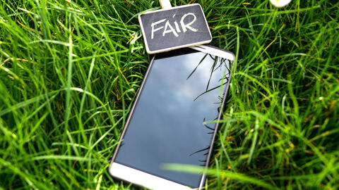 Ein Smartphone liegt in einer grünen Wiese. Mit einer Holzwäscheklammer ist eine kleine Schiefertafel am Smartphone befestigt. Auf der Tafel steht "Fair".