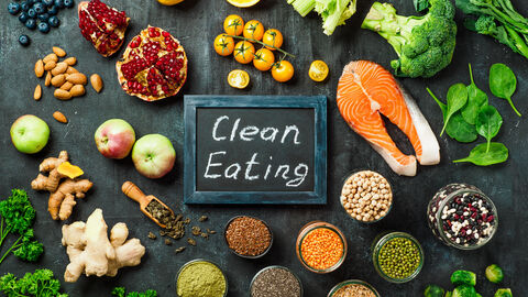 Verschiedenes Obst und Gemüse sowie Nüssel und Lachs liegt um eine Schiefertafel herum. Auf der Tafel steht "Clean Eating". Bei dem Obst und Gemüse handelt es sich zum beispiel um Spinat, Brokkoli, Rosenkohl, Peperoni, Zitronen, Tomaten, Äpfel, Blaubeeren, Erbsen, Ingwer, rote Linsen, Mandeln.