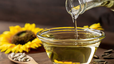 Aus einer Glasflasche wird Sonnenblumenöl in ein Glasschälchen gekippt. Neben dem Glasschälchen liegt ein Holzlöffel auf dem Sonnenblumenkerne liegen. Im Hintergund liegt eine Sonnenblume.