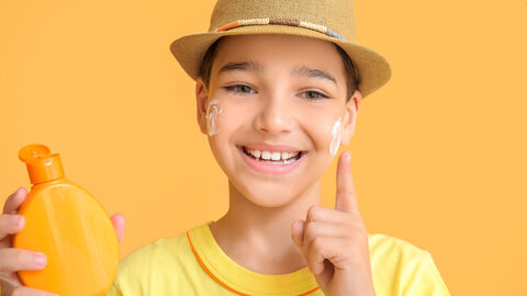 Ein Junge hält mit der rechten Hand eine Sonnencremeflasche in die Kamera. Mit der linken Hand cremt er sich die Wange ein. Er trägt einen Sonnenhut.