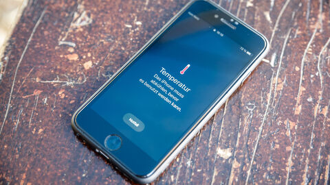 Ein Smartphone liegt auf einem Holztisch in der Sonne. Auf dem Display ist zulesen, dass die Temperatur zu hoch ist und das Smartphone vor der weiteren Nutzung abkühlen muss.