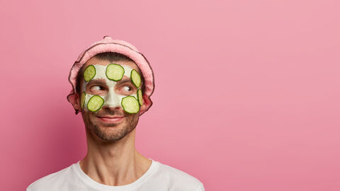 Ein junger Mann trägt eine Creme um die Augen und Wangen. Auf der Creme kleben Gurkenscheiben. Der Mann schaut fröhlich.