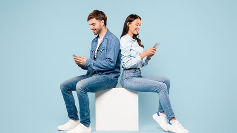 Ein Mann und eine Frau sitzen auf einem Würfel und schauen jeweils auf ihr Smartphone. Der Hintergrund ist einfarbig hellblau.