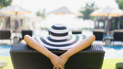 Eine Frau liegt auf einer Liege mit dem Rücken zur Kamera. Es sind nur ihr mit einem Hut bedeckter Kopf und die über die Lehne gelegten Unterarme zu sehen. Im Hintergrund des Bildes ist verschwommen eine Gebäudezenerie mit Pool zu sehen.