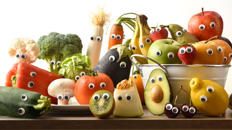 Verschiedene Obst und Gemüsesorten haben aufgeklebte Kulleraugen und sind so hingelegt, dass einen das Obst und Gemüse nun wie für ein Gruppenfoto anschaut. Bei dem Obst und Gemüse handelt es sich beispielsweise um Avocado, Zitrone, Brokkoli, Tomate, Kiwi, Lauch, Zucchini oder Möhren.