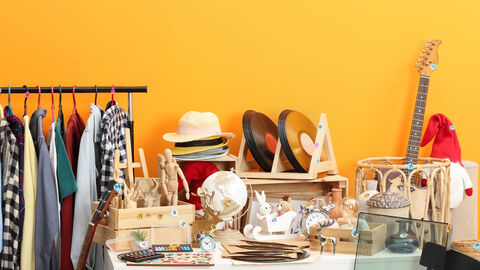 Unterschiedlichste Gegenstände sind an einem Flohmarktstand aufgestellt, wie etwa: Kleidung, Gitarre, Schallplatten, Hüte, Dekoartikel und ein Kindermalkasten und ein Stuhl.