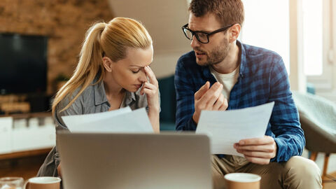 Ein junges Paar sitzt vor einem Computer und hält verschiedene Blätter in der Hand.  Im Hintergrund ist Zimmereinrichtung zu sehen.