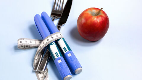 Zwei Abnhemspritzen sind zusammen mit einem Messer und einer Gabel in ein Maßband eingerollt. Daneben liegt ein Apfel.