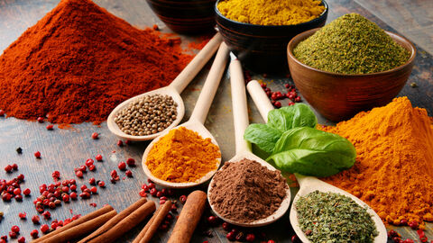 Verschiedene bunte Gewürze auf weißen Löffeln. einige Gewürze sind auch in braunen und weißen Schälchen im Hintergrund. Außerdem gibt es zwei Gewürzhaufen link und rechts im Bild.