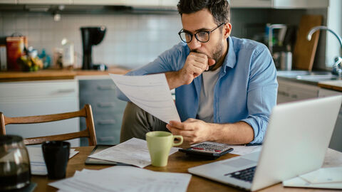 Ein Mann sitzt am Küchentisch. Vor Ihm steht ein Laptop aufgeklappt und es liegen mehrere Zettel vor ihm. Außerdem steht vor ihm eine Kaffeetasse. Er hat ein Bein auf einen Stuhl gestellt und liest mit kristischem Blick eine Rechnung. Der Mann hat dunkle Haare, einen Dreitagebart und hat eine Brille auf.