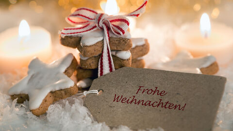 Ein Stapel Zimttserne mit Schleife drm herum steht vor vier Teelichtern. An den Keksstapel gelehnt ist ein Kärtchen mit der Aufschrift "Frohe Weihnachten". Die Kekse und das Kärtchen sind von Kunstschnee umgeben.