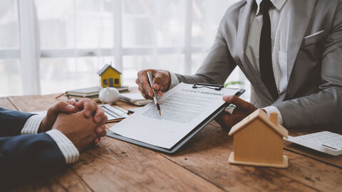 Ein Blick auf einen Schreibtisch. Rechts zeigt ein Mann in einem grauen Anzug mit einem Kugelschreiber auf einen Vertrag auf einem Klemmbrett. Ihm gegenüber sitzt eine weitere Person im Anzug - man sieht allerdings nur die Hände. Auf dem Schreibtisch stehen zwei Häuschen aus Holz.